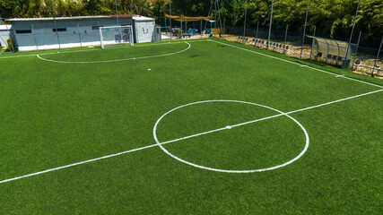 Aerial closeup of a vibrant green synthetic turf soccer field, highlighting the white center circle and midfield line markings, enclosed by a metallic mesh fence. The football pitch is empty.  © Stefano Tammaro