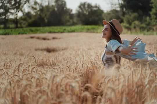 Happy young woman in a hat and blue shirt walking through a golden wheat field at sunset