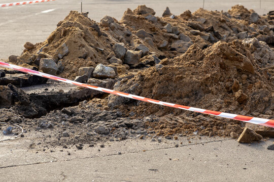 Road construction trench with piles of dug soil and rubble cordoned off by striped red white warning tape, marking an urban utility repair project site for infrastructure maintenance