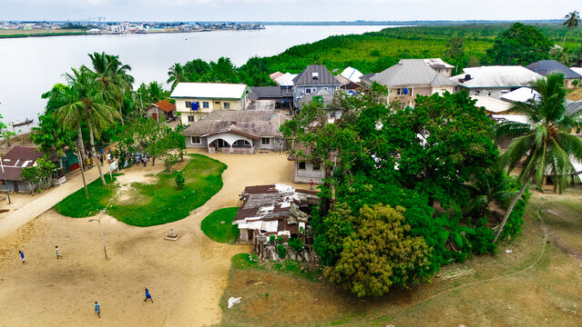 Aerial view of a riverside village with houses, lush trees, and people walking on a sandy path along the river in Minima, Rivers, Nigeria.