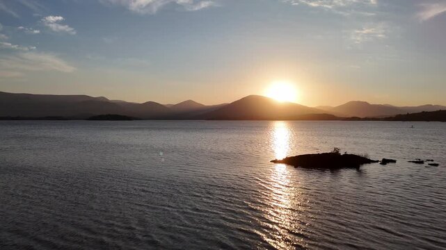 Aerial drone shot of Loch Lomond along the West Highland Way in Scotland. The drone does a pull back revealing the lake with a sunset and mountains in the background.