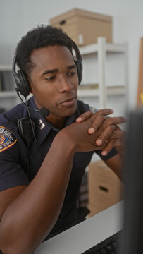 Police officer speaks into headset with hands clasped at a desk in a building office by a computer monitor and visible badge; calm focus.