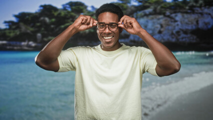 Naklejka premium Man adjusting eyeglasses and smiling in studio while facing camera, hands on frames at temples, wearing casual t shirt and standing before a beach backdrop; joy vacation ease.