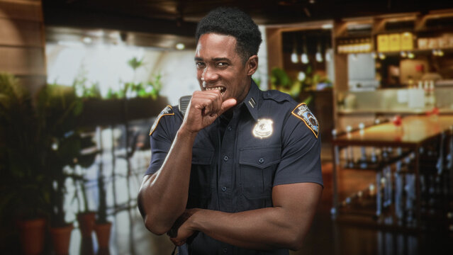 Man police officer holds radio mic and shows badge, smiling in restaurant interior inside a building, short sleeve uniform visible; duty service approachable.