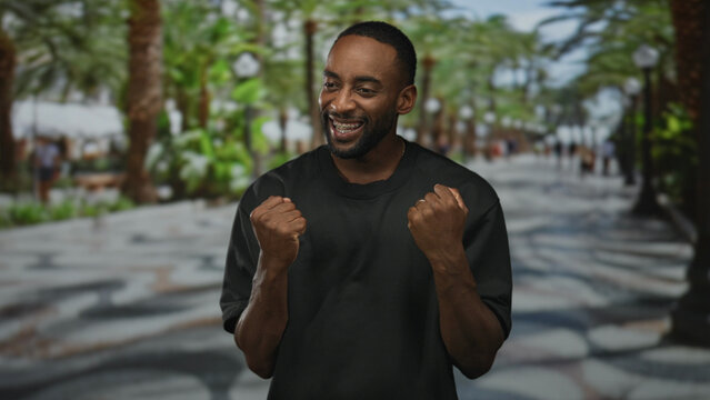 Young african american man with clenched fists and visible braces smiling on busy seaside street promenade with palm trees; triumph.