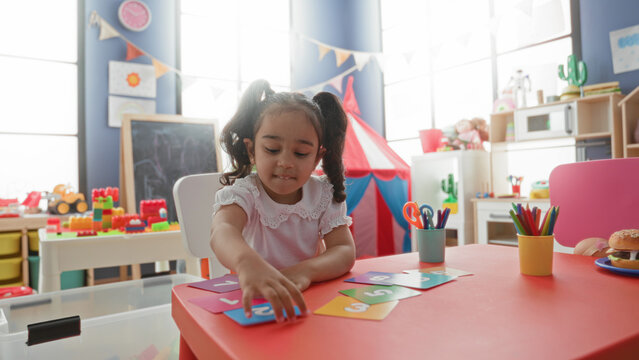Young girl sorting colorful number cards with small hands at a red table in a building playroom with toys and craft cups; concentration learning play.
