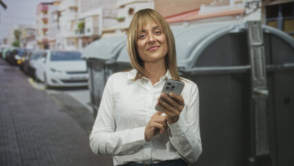 Fototapeta premium Woman tapping smartphone with right hand on urban street by dumpsters and parked cars; everyday connection contentment.