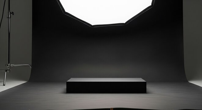 An empty photo studio setup with a black backdrop, a small black display podium, and a bright softbox light overhead