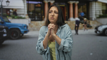 Fototapeta premium Woman with hands clasped pleading on a city street in front of a building entrance, eyes closed and brow furrowed; desperation.