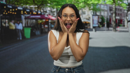 Fototapeta premium Young woman presses hands to cheeks in shocked surprise on street with cafe scene and outdoor lights; spontaneous joy.