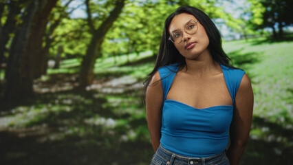 Fototapeta premium Woman with bare shoulders and blue sleeveless top tilts head to side in studio, wearing glasses and hands behind back; serene reflection.