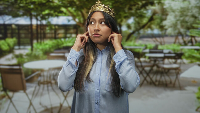 Woman wearing gold crown and light blue shirt holds hands to ears and winks while grimacing on outdoor restaurant terrace with metal tables; annoyed.