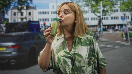 Fototapeta premium Woman holding green juice bottle, sniffing near nose and smiling on a street with cars and cyclists; curiosity wellness.