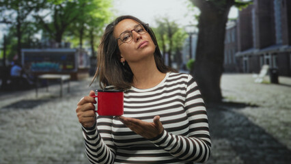Naklejka premium Woman holding a red mug and presenting open palm on a sunlit cobblestone street near benches and a kiosk; skepticism.