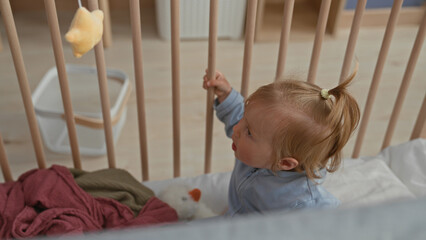Toddler girl sitting in a wooden crib holding a plush duck and touching her sock while looking down in a building  gentle curiosity. © Krakenimages.com