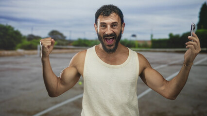 Naklejka premium Man holding smartphone with raised fist and broad smile in a paved street parking area, celebrating a small victory on screen; triumph joy.