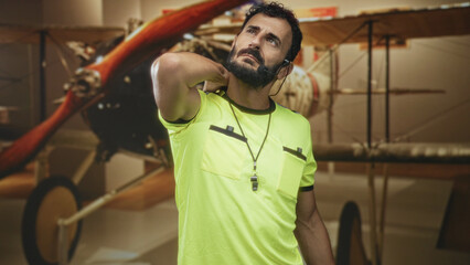 Man referee middle aged bearded hispanic with whistle and headset, hand behind neck stretch in a building with vintage biplane exhibit  tired reflection. © Krakenimages.com