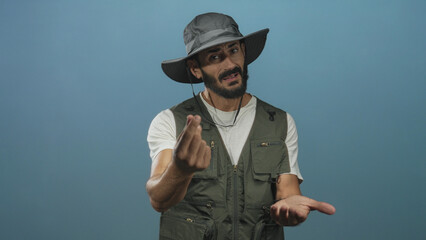 Fototapeta premium Man with beard wearing wide hat and fishing vest beckons with fingers and holds an open hand forward in studio; simple invitation.