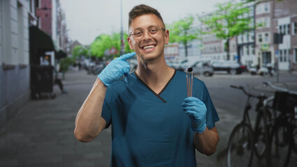 Fototapeta premium Man dentist in blue scrubs wearing blue gloves holds dental mirror and probes while pointing finger to cheek on street, smiling broadly at camera; friendly care.