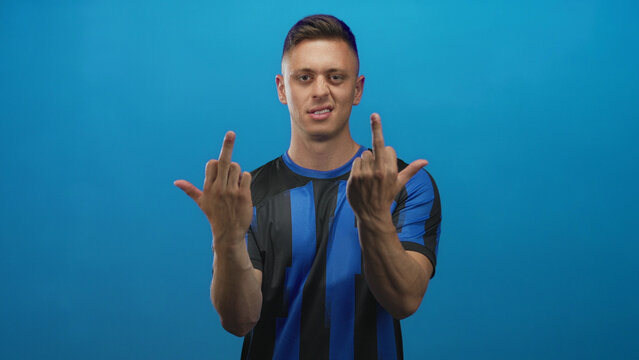 Young blond man in blue and black soccer jersey shows both middle fingers in a studio setting; defiance attitude.