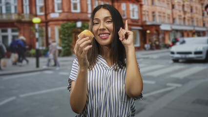 Fototapeta premium Hispanic brunette young woman holding a muffin in one hand points finger up on a city street crosswalk while smiling broadly; joy.