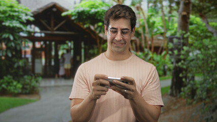 Fototapeta premium Man smiling and looking down at a smartphone, hands holding the phone while standing near a building entrance framed by tropical foliage; contentment.