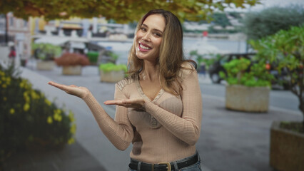 Fototapeta premium Woman palms up presenting with open hands and smiling on a city street beside planters and walkway; friendly welcome.