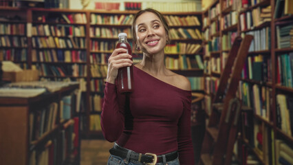 Woman holding a bottle of dark juice in a library building near bookshelves and ladder, casual maroon off shoulder top, jeans and belt visible  playful confidence. © Krakenimages.com