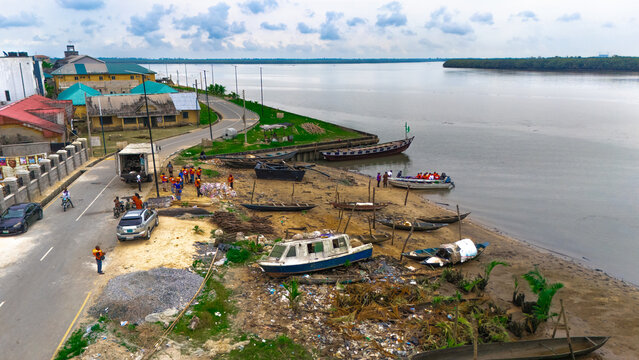 Aerial view of a riverbank with wooden boats, small motorboats, and people gathered near a road, with buildings and vegetation along the shore in Minima, Rivers, Nigeria.