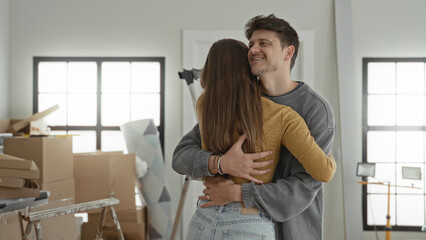 Young man hugging woman with hands clasped on back amid packed moving boxes in building; home joy...