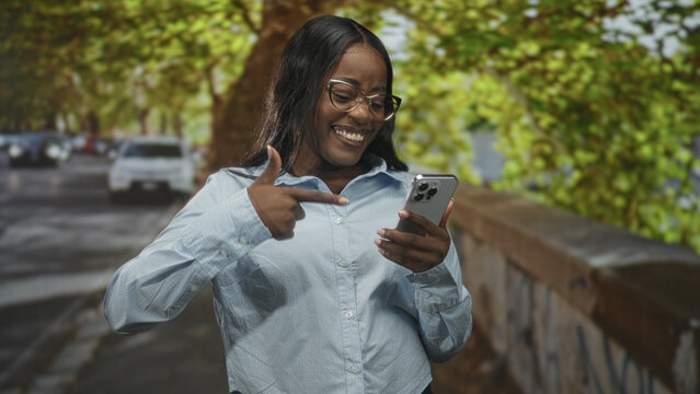 Woman holding smartphone and pointing finger while smiling on a tree lined street beside a stone railing; joy connection.