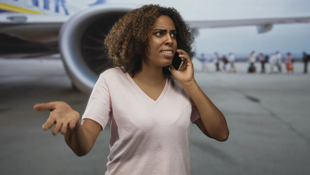Woman talking on phone at airport terminal near plane engine, hand to ear while reaching toward gate with queued passengers; travel delay concern.