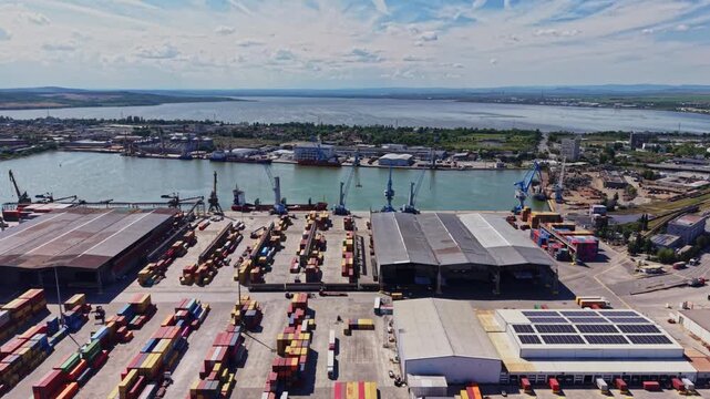 Containers are stacked at a busy shipping yard near a river. Cranes are loading and unloading goods. The scene shows workers and vehicles moving around during daylight.
