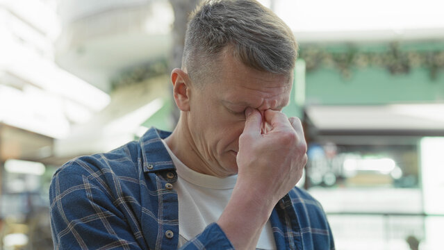 Man pinching bridge of nose and rubbing eyes on a busy urban shopping street in front of storefronts and awnings; fatigue.