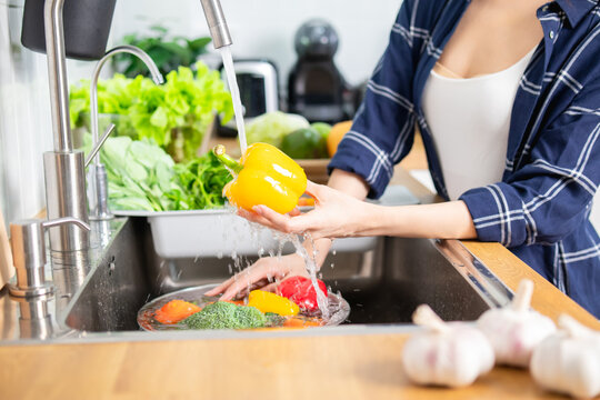 Woman washing fresh red bell pepper under running water in kitchen sink