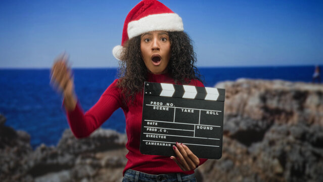 Woman wearing a santa hat holds a clapboard while covering her face with one hand at a studio seaside cliff set; embarrassment holiday fun.