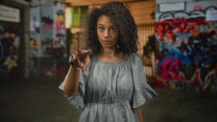 Naklejka premium Teen woman with curly hair pointing finger toward camera on street with graffiti covered wall and building; playful confidence.
