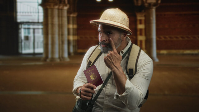 Man holding passport and pointing finger while standing in a historic building hall with backpack and pith helmet; confidence travel exploration.
