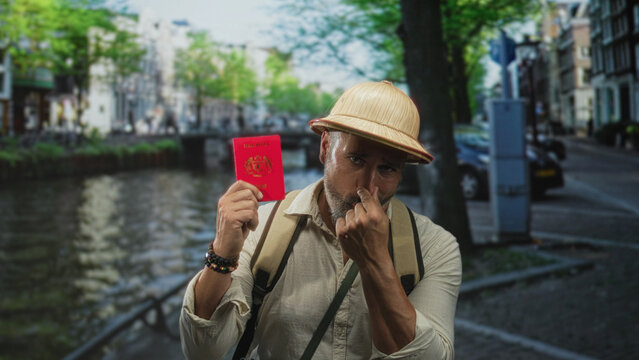 Man holding red passport and touches nose while wearing pith helmet and backpack on street by canal; playful travel adventure.