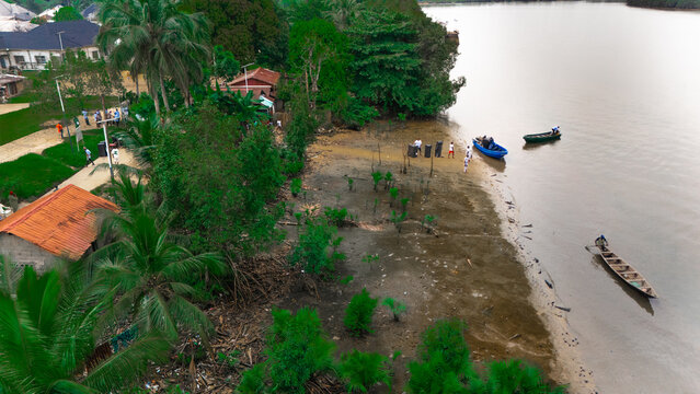 Aerial view of a riverbank with small wooden boats, local people, lush palm trees, and simple houses along the water in Minima, Rivers, Nigeria.