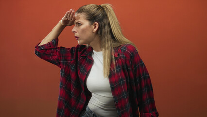 Fototapeta premium Woman in red plaid shirt and white tee shielding eyes with hand while peering to the right, mouth slightly open and ponytail visible in studio; curiosity exploration.
