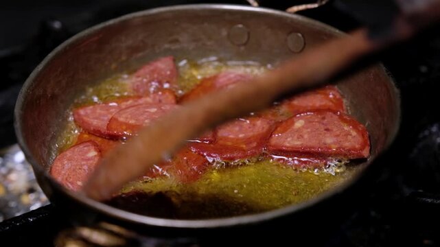 Sucuk slices are cooked in oil in a copper pan. Close-up scene showing hot breakfast food preparation, pan cooking process and appetizing sucuk detail.