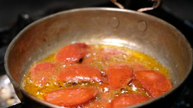 Sucuk slices are cooked in oil in a copper pan. Close-up scene showing hot breakfast food preparation, pan cooking process and appetizing sucuk detail.