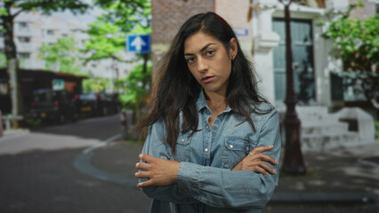 Fototapeta premium Woman arms crossed on city street in denim shirt, hands visible with ring and stern gaze; pensive resilience.