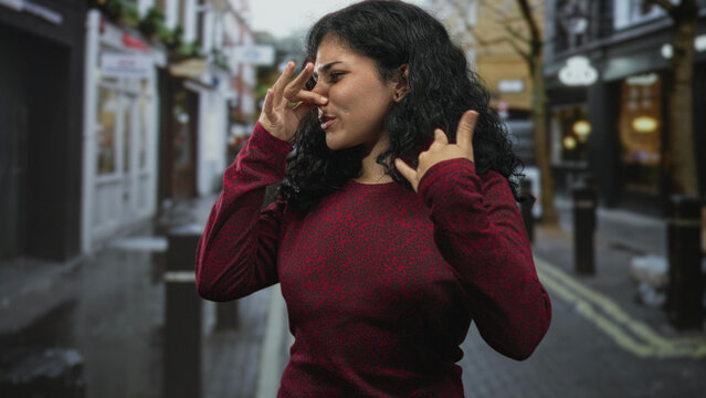 Young arab woman pinching her nose with raised hand on crowded city street, wearing red sweater and wincing at a strong smell; disgust avoidance.