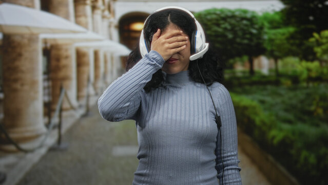 Woman wearing headset with hand covering eyes and palm raised in stop gesture on a stone colonnade street walkway; frustration.