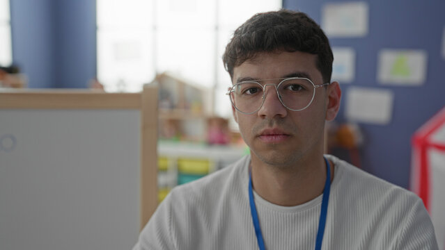 Young man with glasses and blue lanyard looking sideways near a whiteboard and playhouse in a preschool classroom; calm focus patience care.