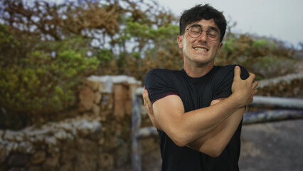 Fototapeta premium Man smiling with arms crossed hugging himself in a forest setting by stone wall, wearing glasses and black shirt; comfort selfcare.