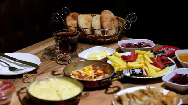 A rich Turkish breakfast is served on a wooden table with kuymak, sucuklu eggs, French fries, bread, tea, jam, honey, cheese and assorted breakfast dishes.