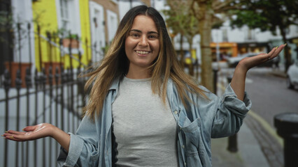 Fototapeta premium Woman smiling with palms up and open hands on a street near a yellow building; joy playful openness.